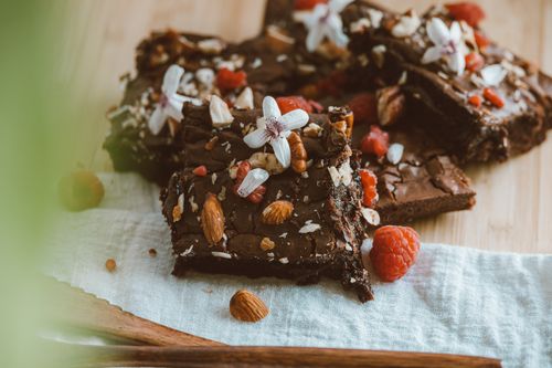 Free Close-up of chocolate brownies adorned with almonds, raspberries, and white flowers on a wooden board. Stock Photo
