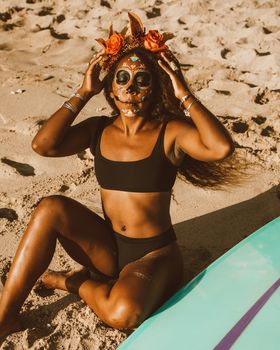 Woman in swimwear with Day of the Dead makeup sitting on sandy beach next to a surfboard.