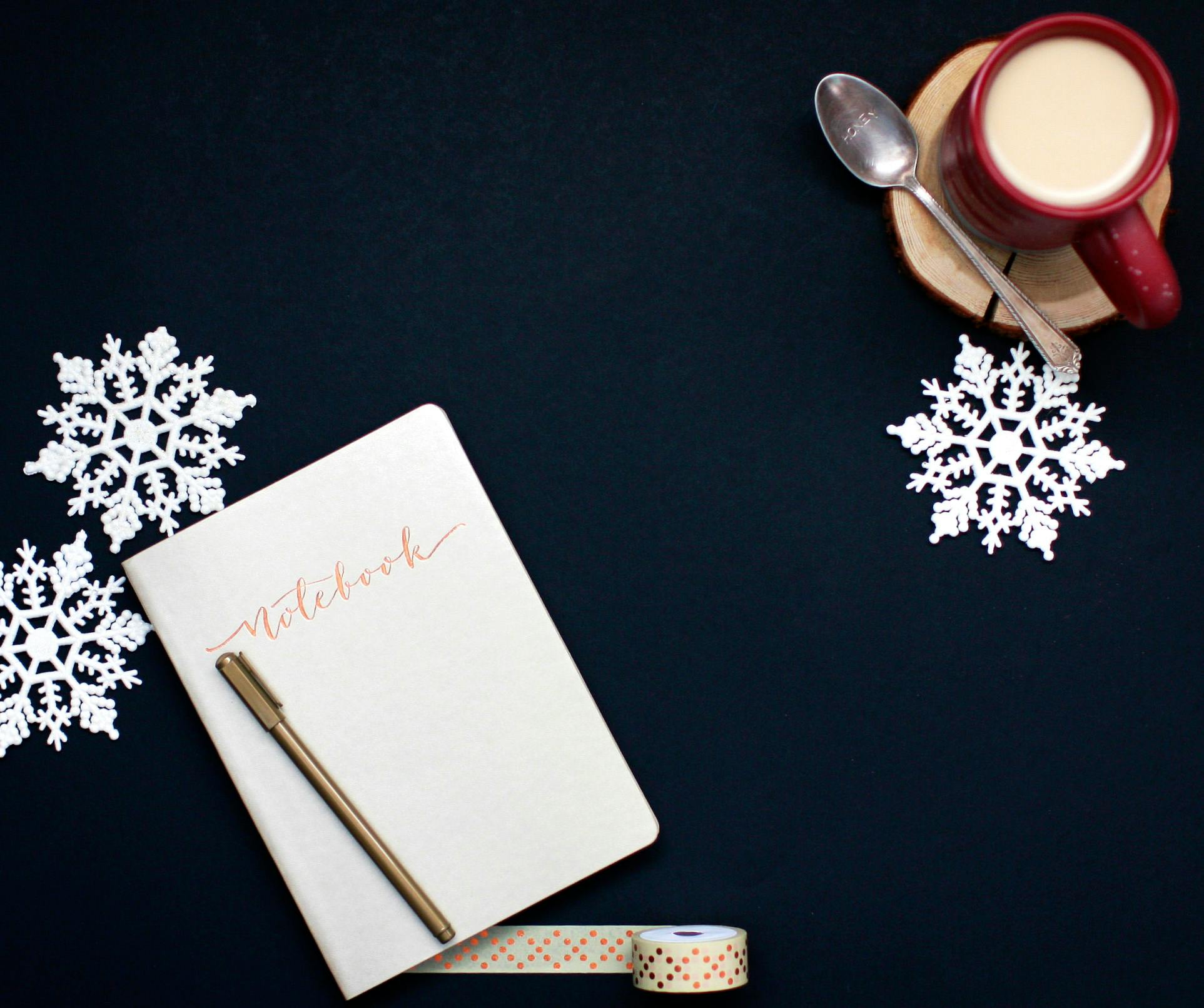 brown pen on white notebook near red coffee mug and silver spoon on top of blue surface