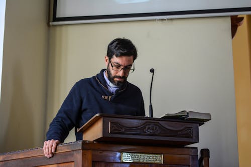Serious bearded male teacher in casual clothes and eyeglasses reading lecture while standing at wooden podium in conference hall
