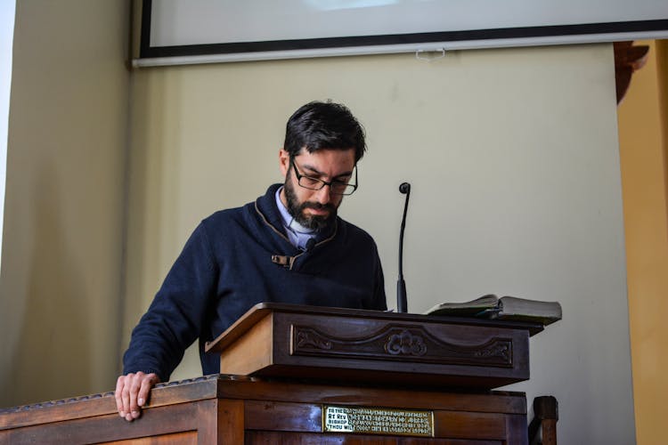 Focused Male Lecturer Reading Report On Tribune With Microphone