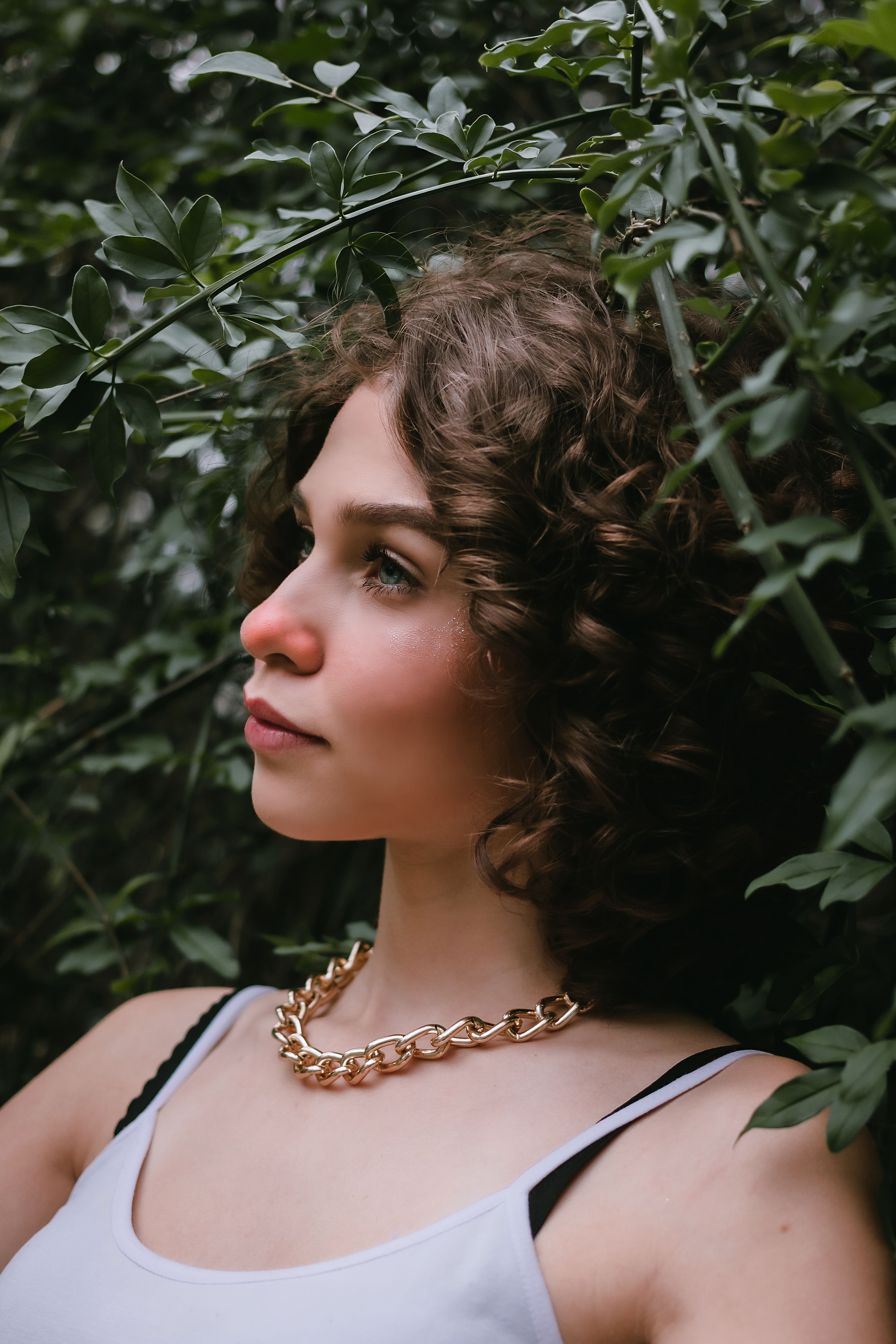 Dreamy young female model with curly hair in casual clothes and golden necklace standing near lush green plants in garden