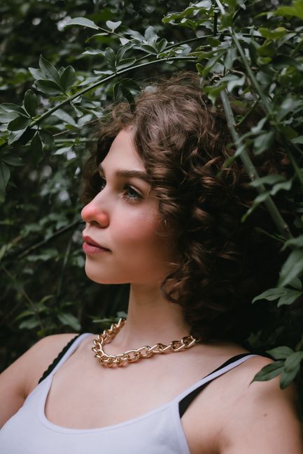 Dreamy young female model with curly hair in casual clothes and golden necklace standing near lush green plants in garden