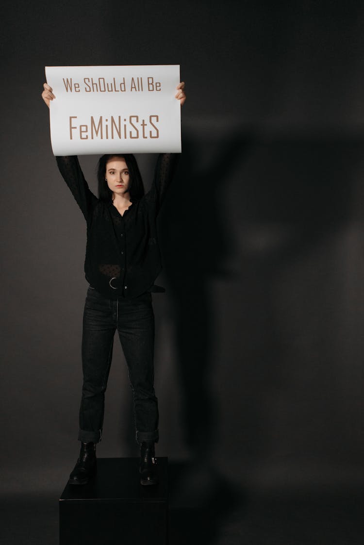 A Woman In Black Long Sleeves Standing On A Block While Holding A Poster