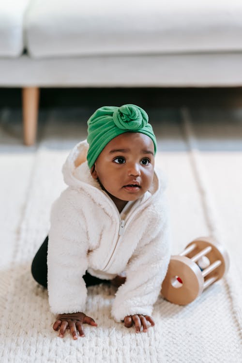 Curious adorable infant Indian girl wearing green turban crawling around white carpet with wooden sensory toy and looking away