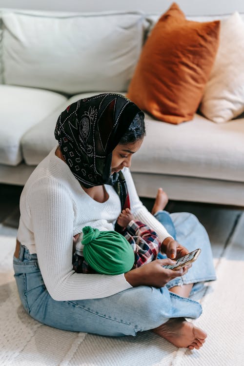 Free Side view of young Indian female in casual clothes and headscarf embracing newborn baby and messaging on mobile phone while sitting on floor in living room Stock Photo