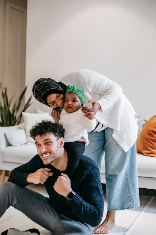 Adorable little Indian girl sitting on shoulders of happy father while playing together with mother in cozy living room