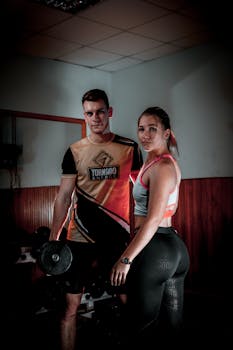 A muscular man and woman posing with dumbbells in a gym setting, showcasing fitness and strength.