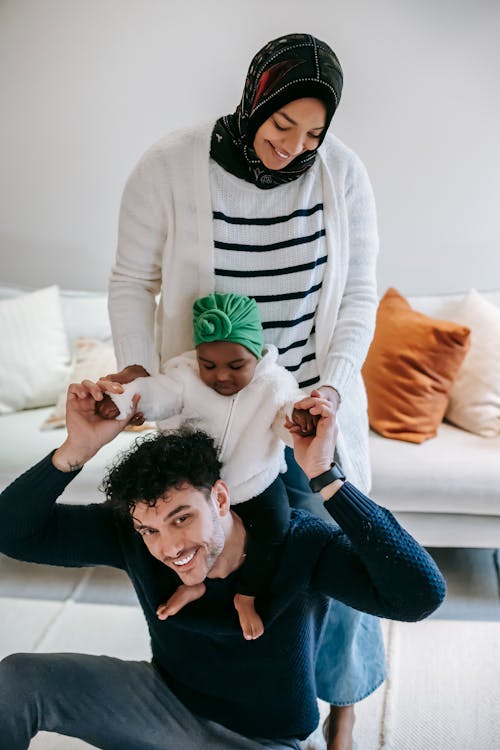 Positive father with black daughter on shoulders supported by mother standing behind in living room against sofa