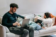 Focused dad using netbook sitting near Muslim mom feeding baby on sofa in living room