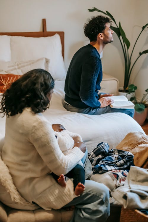 Free Side view of calm African American mother with newborn baby sitting near ethnic father on bed with book while spending time together at home Stock Photo