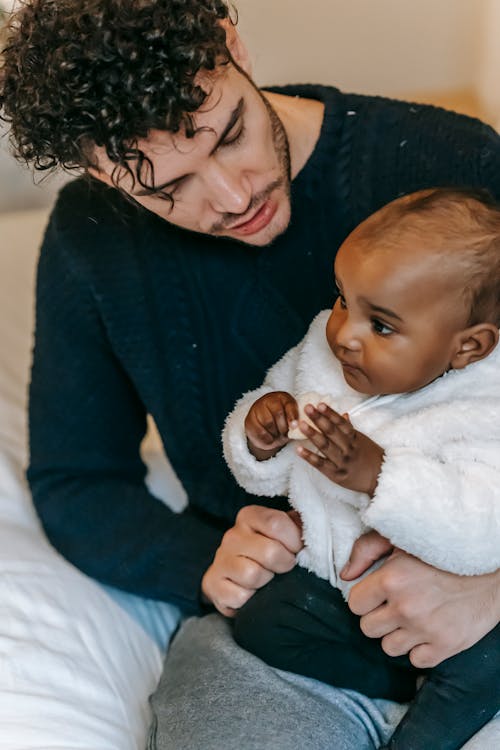 Free Caring ethnic man with beard and curly hair playing with African American infant baby while sitting on bed in bedroom Stock Photo