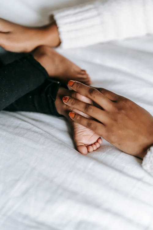 Free Unrecognizable African American mother touching barefoot foot of faceless infant baby while lying on white blanket in bedroom on bed Stock Photo