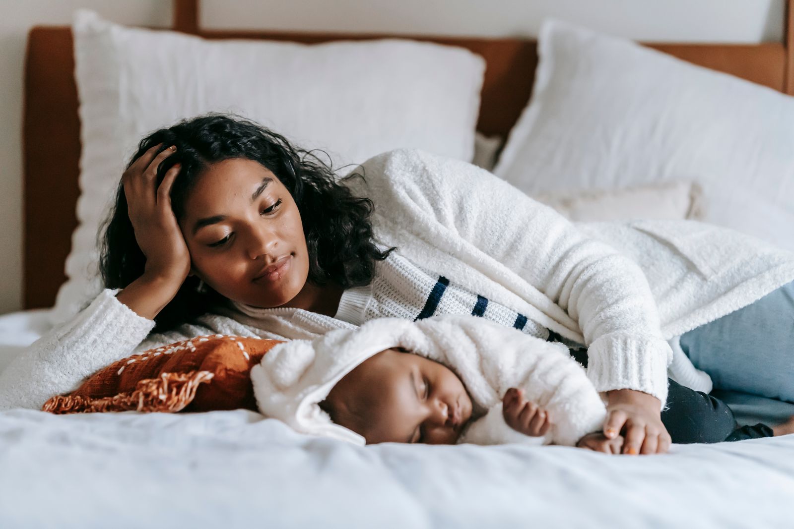 A Black mother reclines on a bed beside her sleeping Black infant, head propped on her hand, gazing thoughtfully at the baby in a sunlit domestic interior.