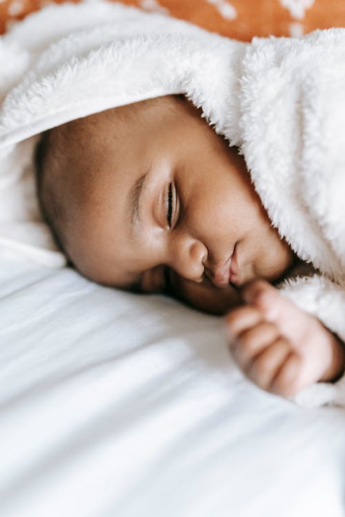 High angle of sweet newborn ethnic baby in soft warm pajama sleeping on cozy bed in sunlight