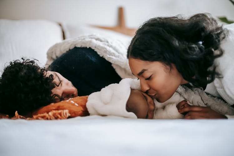 Young Ethnic Mother Kissing Baby Sleeping Near Dad On Bed