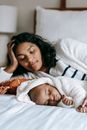 Tranquil young ethnic female in casual clothes lying on bed near cute sleeping baby in sunny morning at home