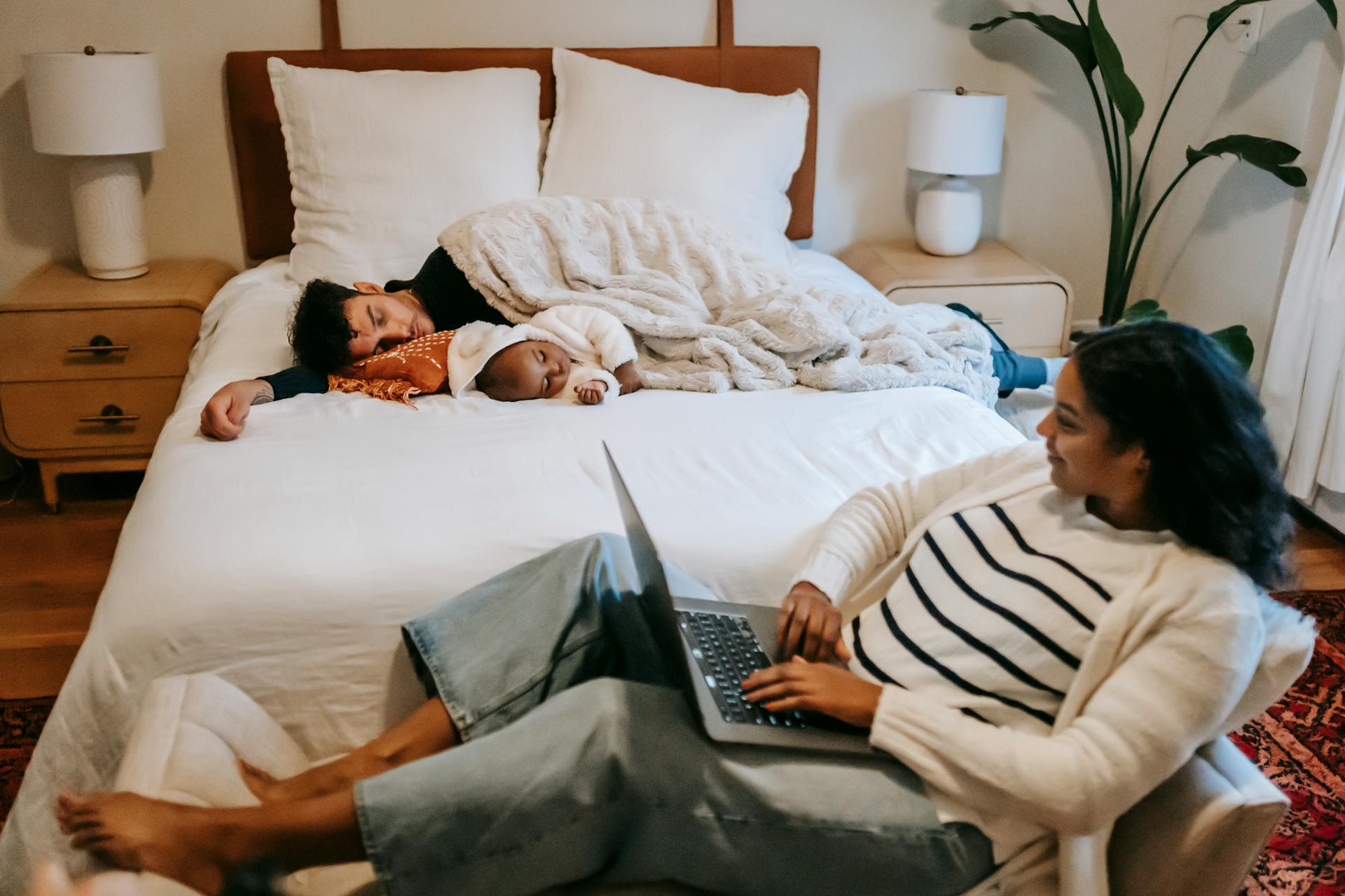 A mother works on a laptop while her family rests peacefully on a bed in a cozy bedroom.