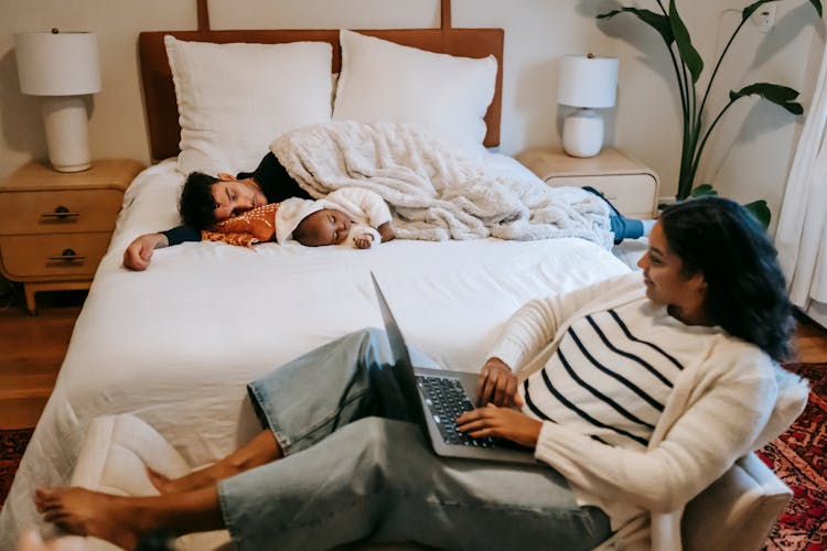 Young Ethnic Woman Using Laptop Near Husband And Baby Sleeping On Bed