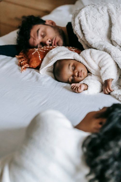 Free From above of unrecognizable ethnic female lying in bed and looking at sleeping infant child and husband during weekend at home Stock Photo