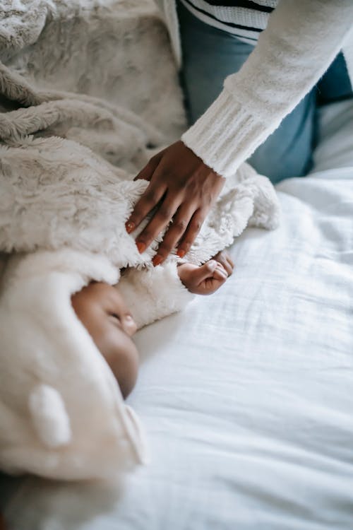Free From above of crop unrecognizable African American mother in casual clothes covering cute newborn baby sleeping on bed with soft plaid in sunny morning Stock Photo