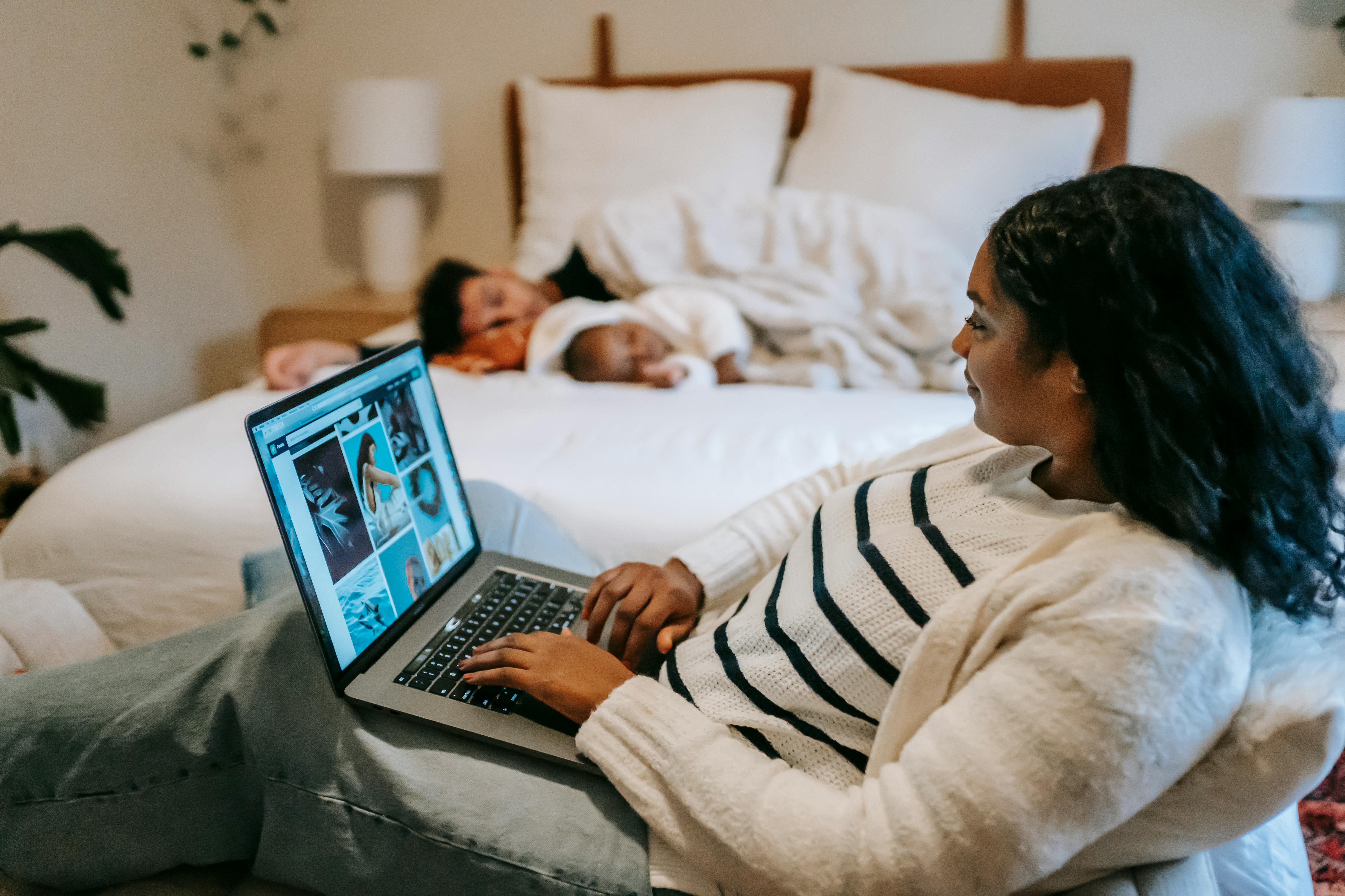 ethnic mother using laptop near baby sleeping on bed with father