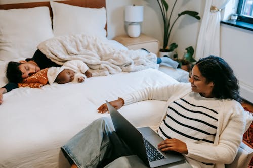 High angle of joyful young ethnic mother smiling while watching movie on netbook sitting near adorable baby sleeping on bed with father during weekend at home