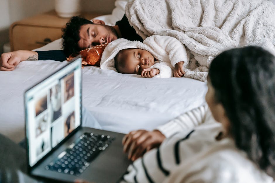 Anonymous ethnic mother working on netbook near baby and husband sleeping in bedroom