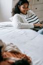 Crop smiling African American female typing on laptop and looking at unrecognizable husband sleeping on bed in daylight