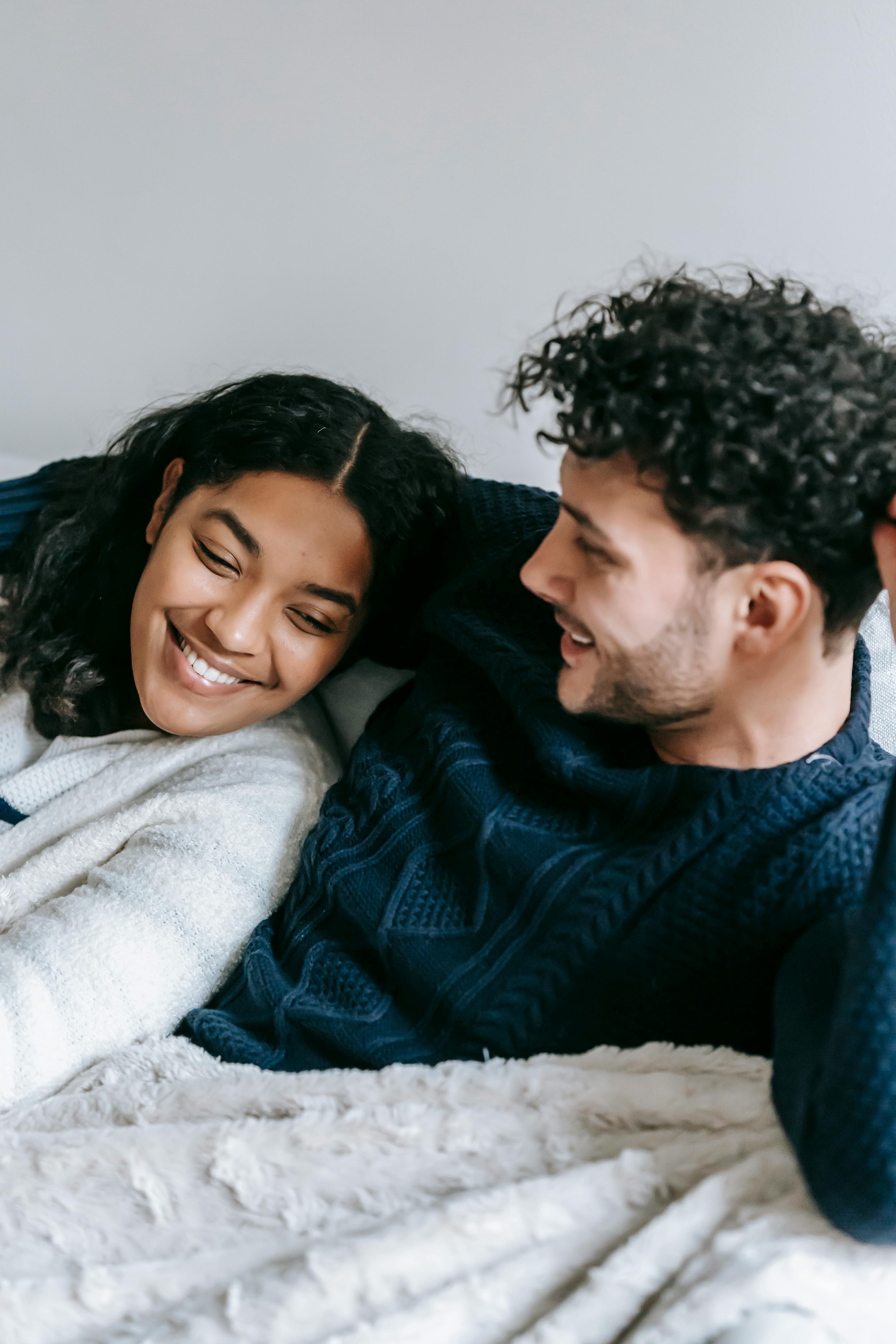 Happy young man embracing smiling African American girlfriend while relaxing together on cozy sofa during lazy weekend at home