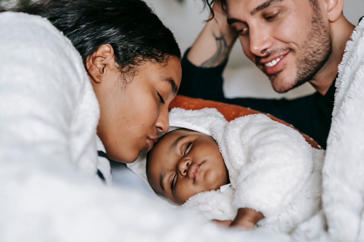 High angle of crop young African American woman kissing sleeping cute newborn baby while resting in bed with happy husband