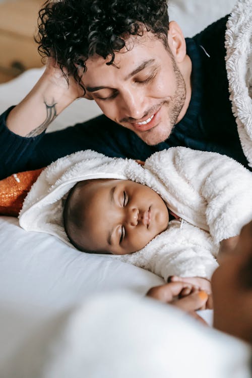 Smiling ethnic father looking at cute sleeping African American newborn baby while lying on bed in cozy bedroom at home