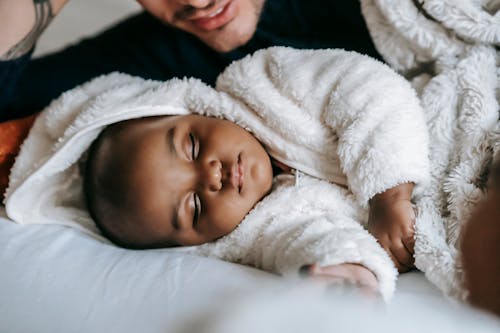 Unrecognizable ethnic father lying on bed with African American newborn baby in white pajama sleeping in bedroom during bed time at home