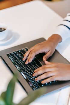 High angle of crop unrecognizable ethnic female freelancer typing information on laptop at white table near cup of coffee on saucer near green plant in light workspace