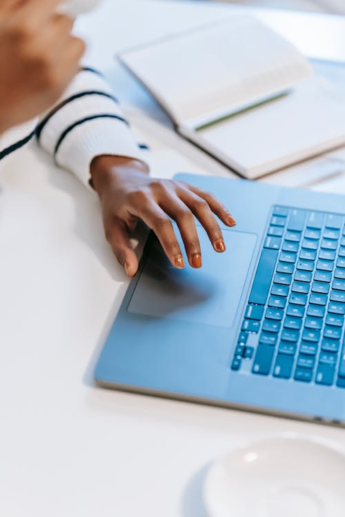 Free High angle of crop anonymous ethnic woman working remotely at white table on laptop near notebook with pen while drinking coffee in light workplace Stock Photo