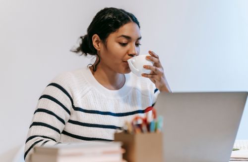 Free Concentrated young Indian female freelancer in casual outfit using netbook at table while drinking coffee near holder for office supplies and books in light workspace Stock Photo