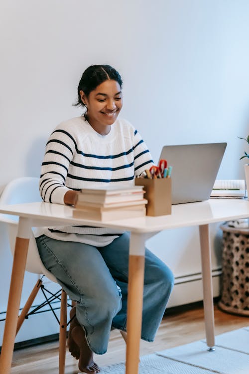 Full length of happy young ethnic female freelancer in casual outfit sitting in chair at table while browsing on netbook near books and holder with office supplies in bright workspace