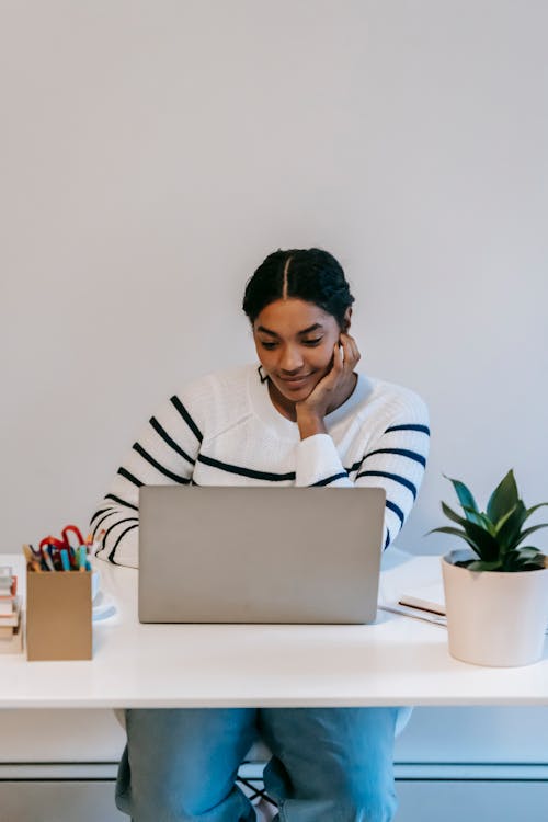 Concentrated young Indian female freelancer in casual outfit using netbook at table near potted green plant and holder for office supplies in bright workspace