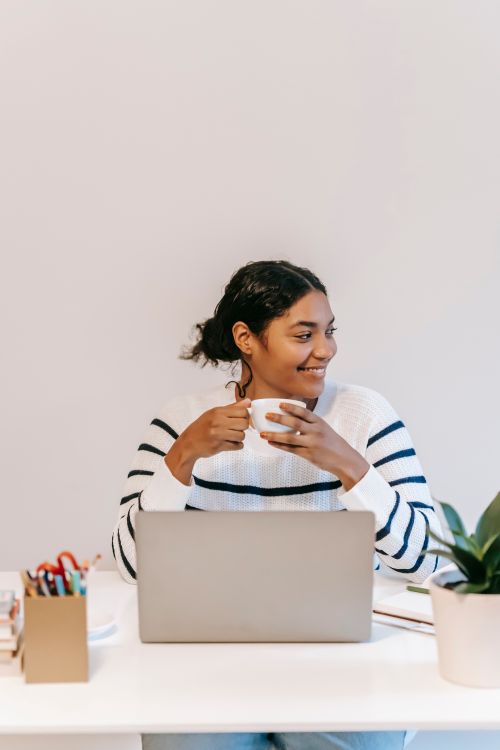 Free Positive ethnic lady in casual clothes working remotely on netbook at table while drinking tea near potted green plant and holder for office supplies while looking away in room Stock Photo