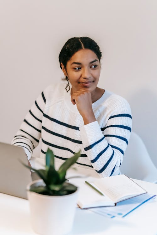 Dreamy young Indian female student in casual clothes sitting on chair at table and surfing on laptop near notepad with pen and potted plant in bright workspace and looking away