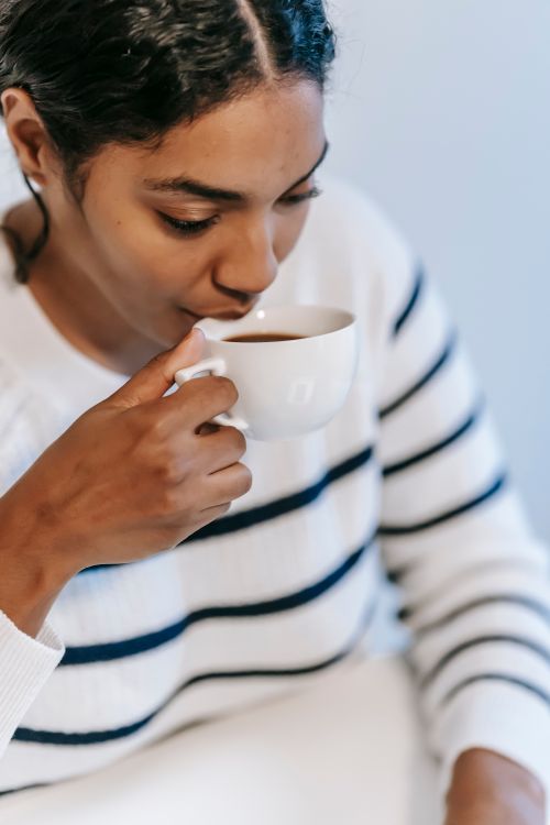 Free High angle of young ethnic lady in casual outfit drinking coffee from white cup at table in light room Stock Photo