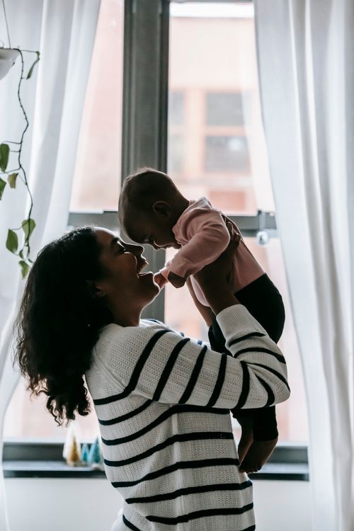Free Side view of positive ethnic mother holding toddler in arms while wearing casual clothes near window with white curtains under potted plant with green leaves in room in daytime Stock Photo