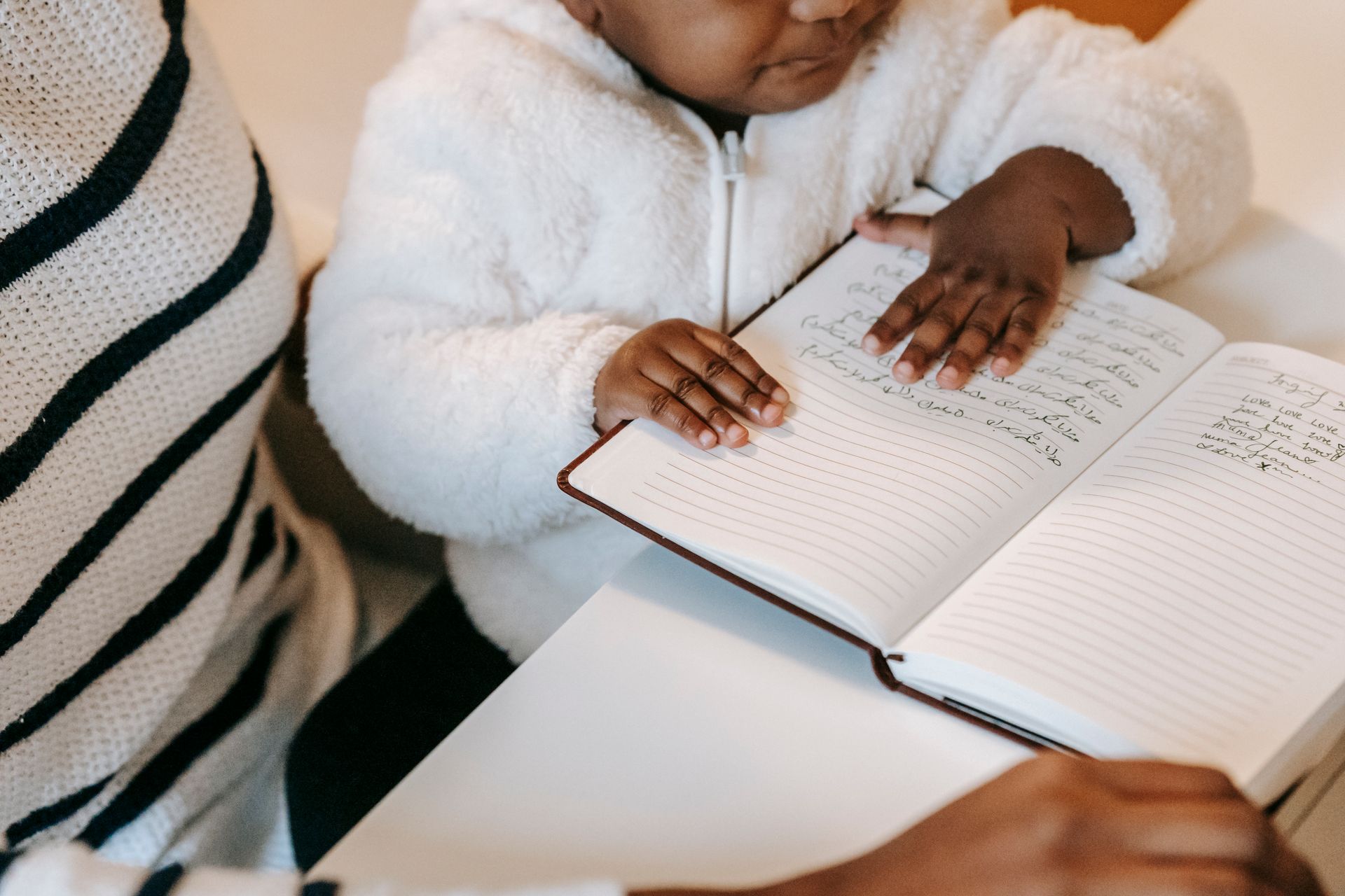 High angle of crop unrecognizable ethnic curious baby examining notepad near lady in casual outfit at table in bright apartment