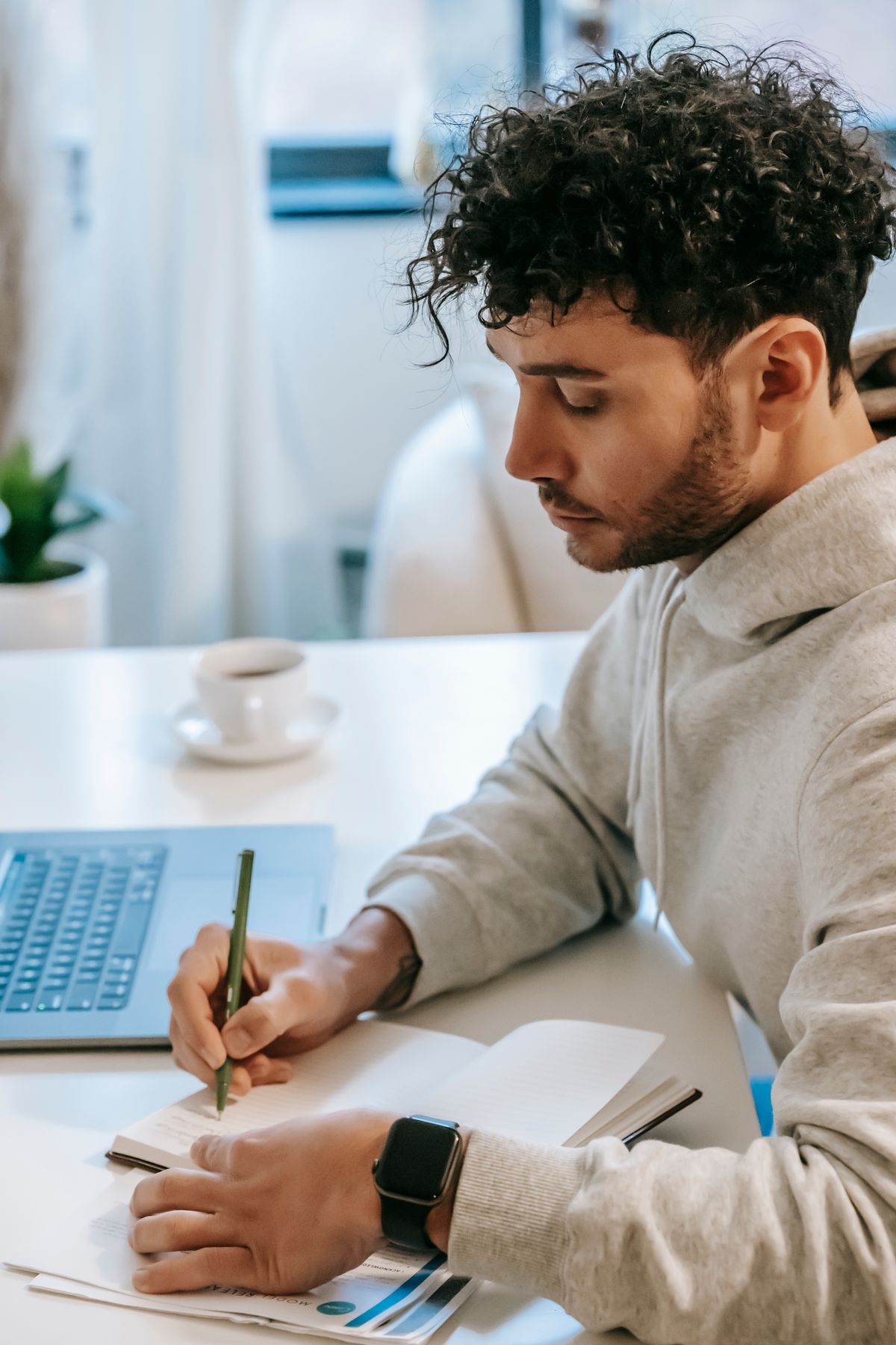Person writing notes in a notebook at a desk beside a laptop and coffee cup