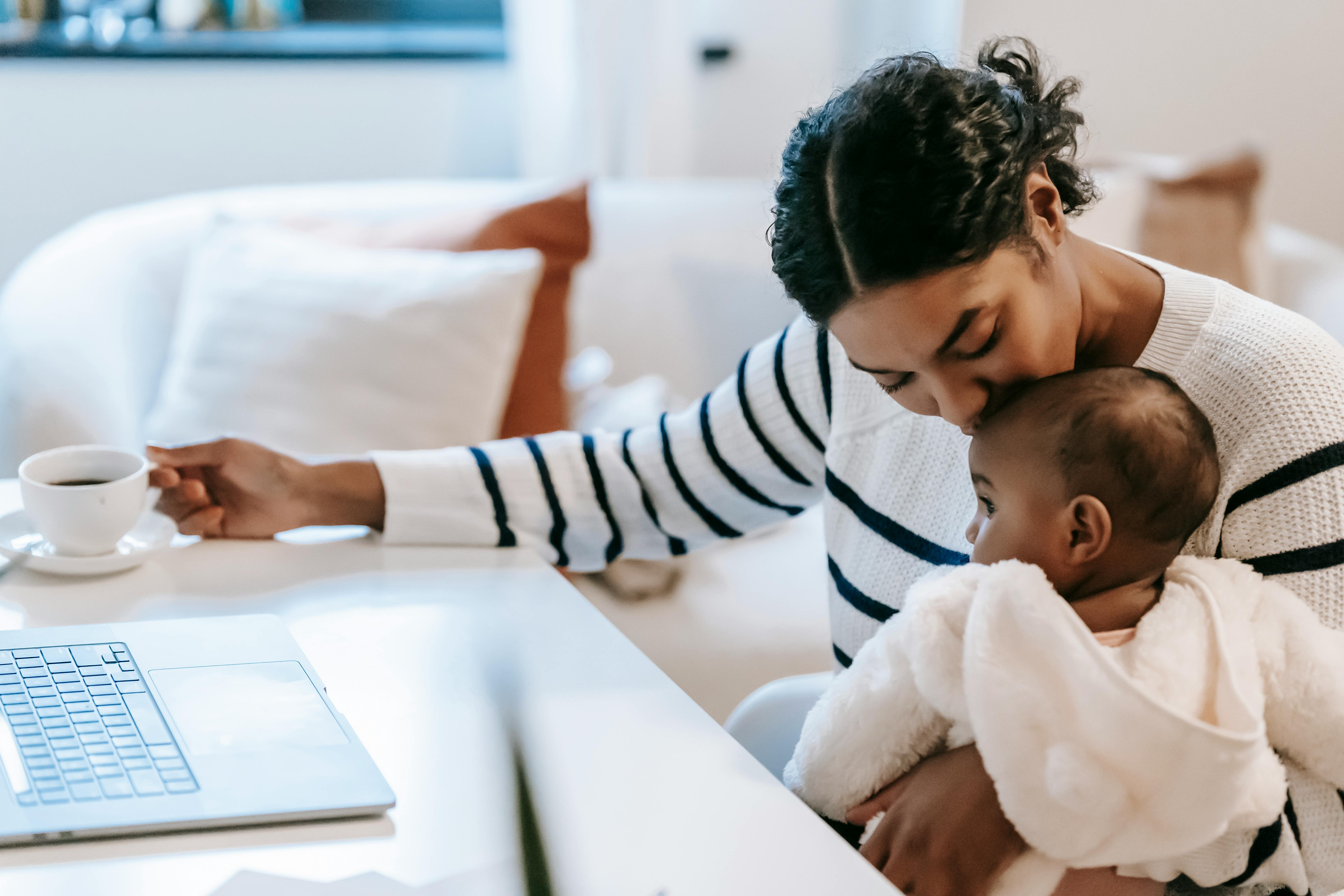 ethnic woman kissing baby near netbook and coffee cup