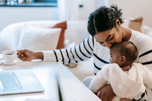 Ethnic woman kissing baby near netbook and coffee cup