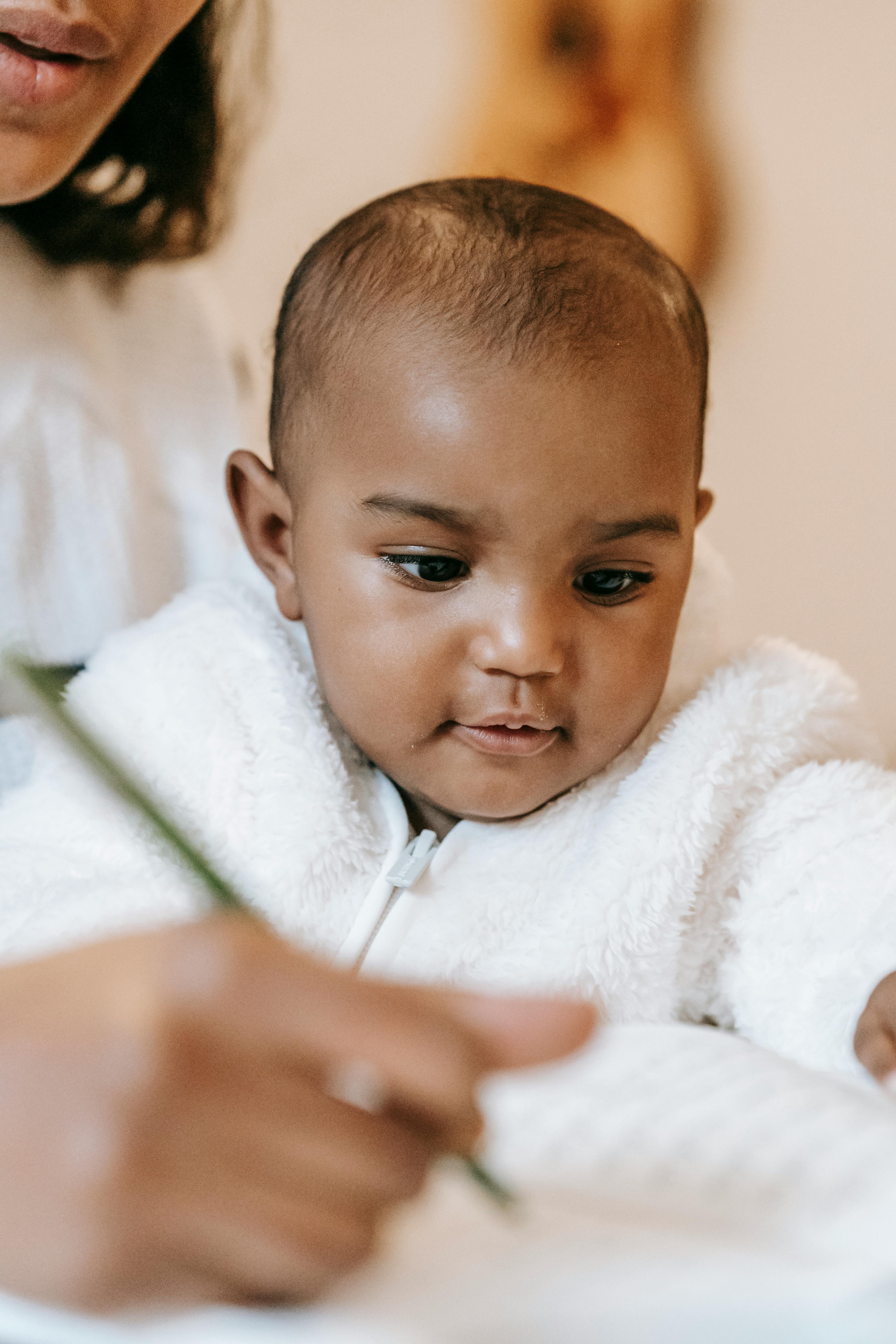 Cute ethnic curious toddler near woman in casual outfit writing notes in notepad with pen at table in light apartment