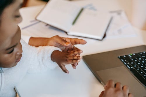 High angle of crop faceless ethnic toddler touching female hand in casual clothes and using laptop at table near opened notepad with pen in light room