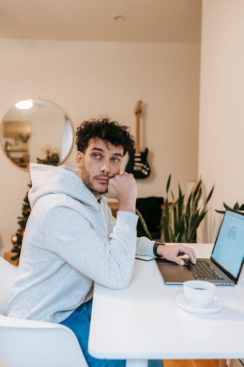 Side view of thoughtful freelancer in casual clothes sitting at table with cup of coffee and laptop while working on project at home in daytime