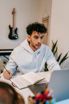 Young bearded male artist writing lyrics in agenda while surfing internet on portable computer in house